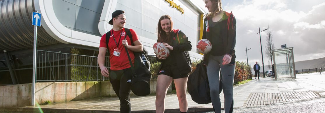 sport 3 students holding rugby balls and sports kit in front of sports building