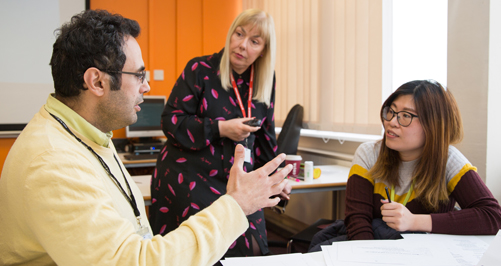 INTL_Courses3 tutor watching 2 learners have a discussion in a classroom